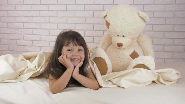 A child with a big toy. Happy little girl on the bed with a big bear.