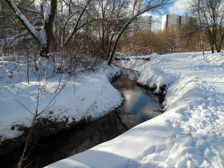 Nonfreezing Ramenka river on a tranquil clear winter day. Moscow. Russia. Fluffy snow adorns leafless trees with festive dresses. Winter nature delights eyes with blinding purity.