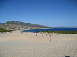 Playa con vegetación, arena y comienzo de dunas. Montañas al fondo.
