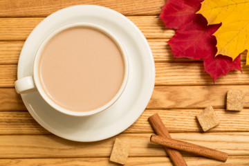 Autumn (fall) cup of tea with milk, cinnamon sticks, brown sugar and colorful bright orange, red, yellow leaves, top view on wooden background, top view.