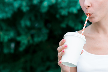 Beautiful young girl stands in a forest with a mug with a red straw. Female hand holding a blank paper cup in a wooded landscape background. Model sucks close up. Mockup for products