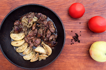Liver, fried with apples and onions in a pan, next to a wooden background whole Apple, black pepper and tomatoes.