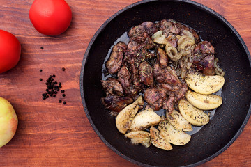Chicken liver, stewed with slices of apples and dried herbs in a frying pan, next to black pepper, tomatoes and a whole Apple, wooden background.