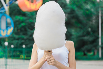 Cute model with cotton candy having fun in the park. Happy girl eating sweet treat bringing back childhood memories. Woman playfully hides her face under cotton candy