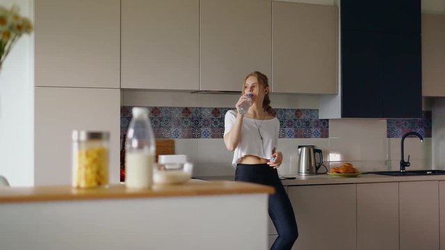 Athletic girl in t-shirt and fitness leggings drinking water from bottle. Young woman in fashion sportswear relaxing in kitchen. Beautiful girl listening music in headphones. After morning run