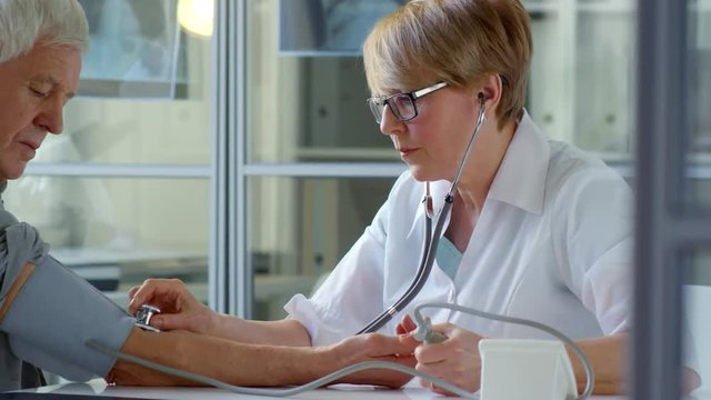Tracking shot of middle-aged female doctor using stethoscope while measuring blood pressure of elderly patient during medical exam