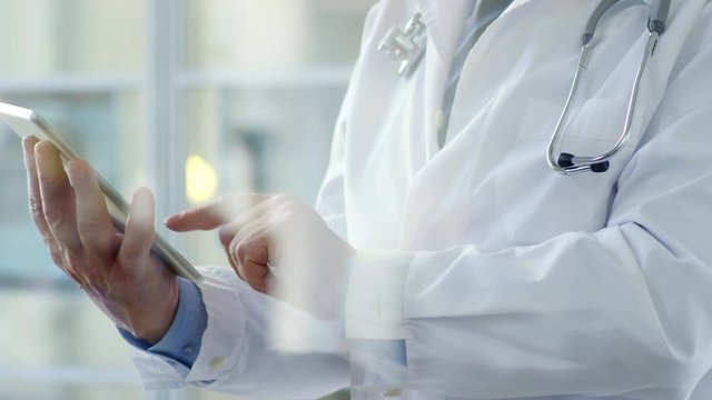 Close-up of unrecognizable male doctor in lab coat with stethoscope over neck standing in office and using digital tablet