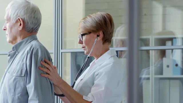 Middle-aged Female Doctor Listening To Back Of Elderly Patient With Stethoscope During Physical Exam In Clinic