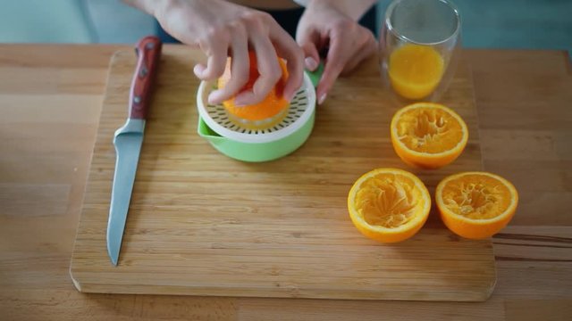 Woman hands making natural orange juice on wooden board. Squeezed orange parts on wooden board. Top view preparing healthy breakfast. Close up female hand rotating part of orange on citrus squeezer