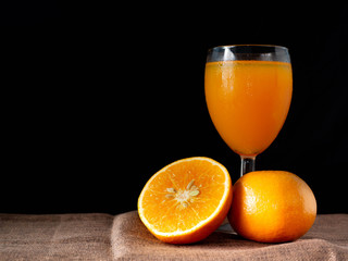Half oranges are placed on a sack on the table, with glass of orange juice on the side on a black background, stilllife