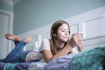 Pre-adolescent teen girl texting on a smartphone lying in bed at home. Candid indoor photo...