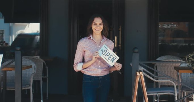 Beautiful mid 30s Caucasian female standing with an open sign near her small cafe, smiling and looking into camera. Small business concept. 4K UHD 60 FPS SLOW MOTION