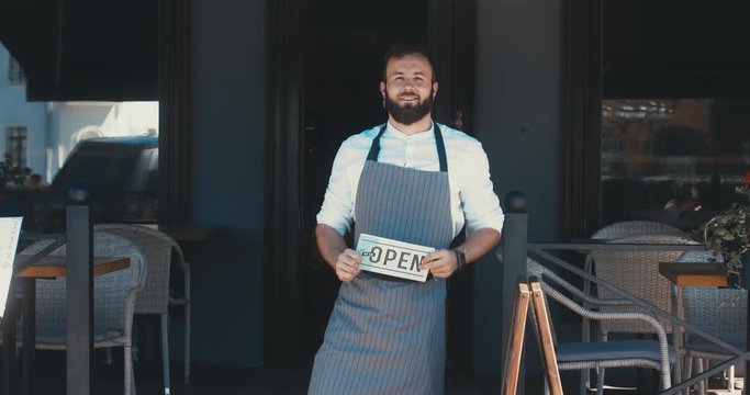 Mid 30s bearded Caucasian male standing with an open sign near entrance of his small cafe, smiling and looking into camera. Small business concept. 4K UHD 60 FPS SLOW MOTION