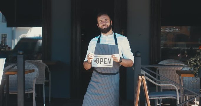 Mid 30s bearded Caucasian male standing with an open sign near entrance of his small cafe, smiling and looking into camera. Small business concept. 4K UHD 60 FPS SLOW MOTION