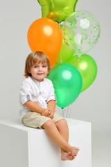 Fototapeta na wymiar Beautiful 3 year old boy with long hair in shirt on white background with bright balloons sitting on the cube