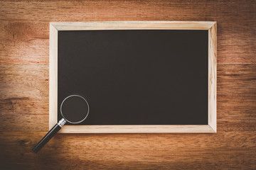 magnifying glass place on rectangle blank slate chalk board with wooden frame on wooden table,...