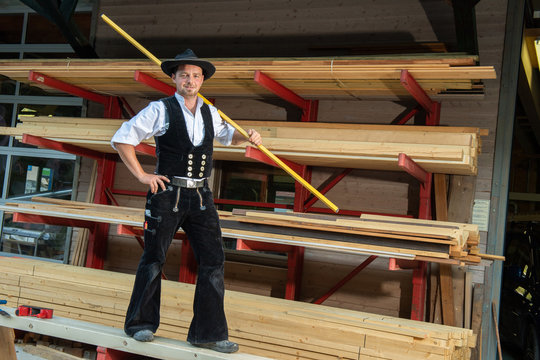Zimmermann in Arbeits Kluft und Schlapphut, mit Wasserwaage über der Schulter, steht auf Holzbalken vor Holzregal