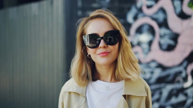 Portrait of beautiful blond girl in sunglasses looking at camera, laughing and touching her hair standing outdoors. Youth, fashion and emotions concept.