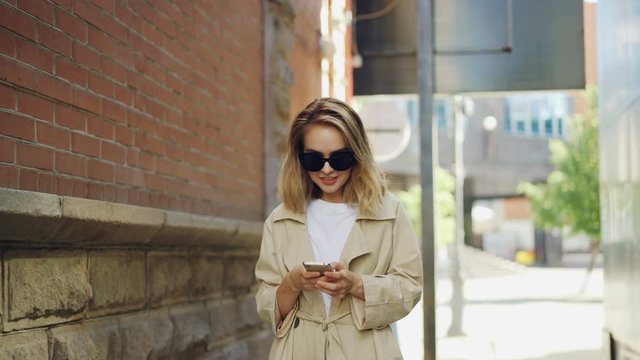 Attractive young woman with blond hair wearing trendy coat and sun glasses is using smart phone walking in modern city. People, modern technology and youth concept.