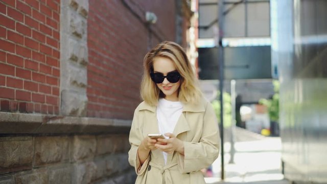 Pretty blond girl is using modern smartphone touching screen walking in city and smiling. Young woman is wearing fashionable clothing and trendy sun glasses.