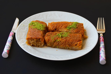 Siyah zemin üzerinde beyaz tabak içerisinde üzeri fıstıklı , kadayıf tatlısı 