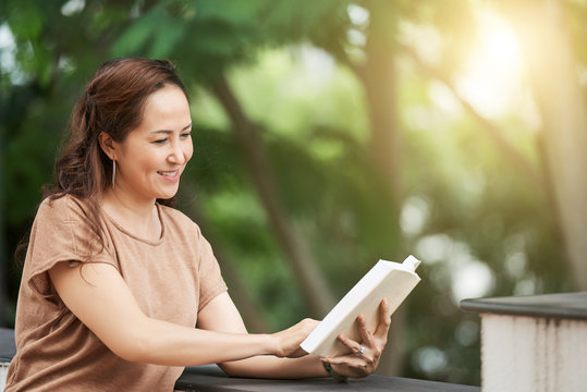 Mature Asian Woman Enjoying Reading A Book Outdoors