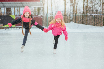 Two Little smiling girls skating on ice in pink wear and hand made scarfs. Outdoor. Winter