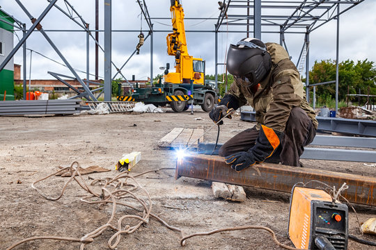 A strong man is a welder in brown uniform, welding mask and welders leathers, a metal product is welded with a arc welding machine at the construction site, blue sparks fly to the sides