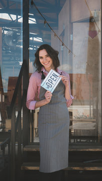 Beautiful mid 30s Caucasian female standing with an open sign near her small cafe, smiling and looking into camera. Small business concept