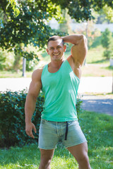 A male bodybuilder stands in a green t-shirt and denim shorts and smiles at the camera