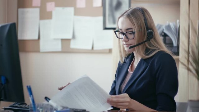 Call center employee at work in the office. A young girl with blond hair with glasses sitting at a table in the office and working with documents. On the head headphones in the hands of a pen and