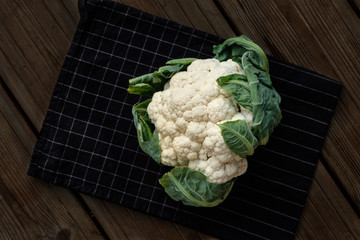 a head of cauliflower with leaves lies on a black in white cage towel on a dark wooden background....
