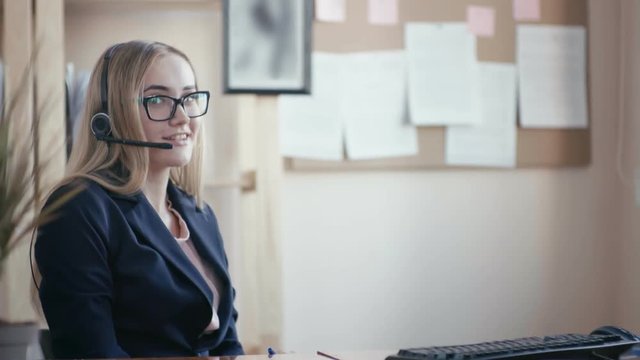 Call center employee at work in the office. A young girl with blond hair with glasses sitting at a table in the office and working with documents. She takes a phone call, holds an automatic pen in her