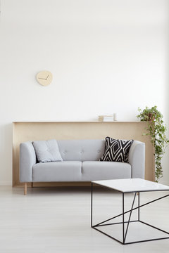 White table in front of grey couch with pillows in living room interior with plant and clock. Real photo