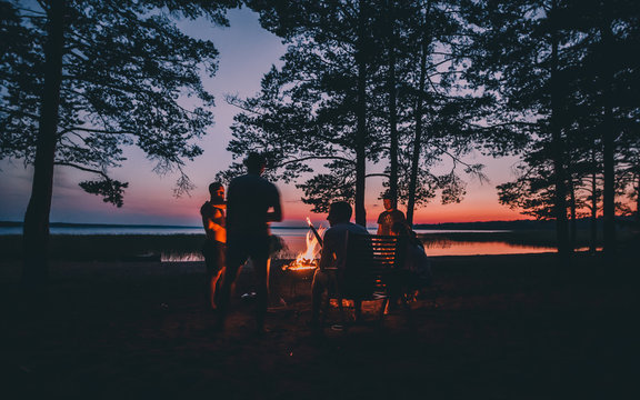 Holiday with friends in nature. Group of young people near camp fire , telling stories near the fire with wood, flames in the nature at night near lake.