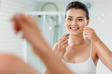 smiling young woman holding dental floss and looking at mirror in bathroom