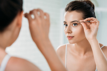 attractive girl correcting eyebrows with tweezers and looking at mirror in bathroom