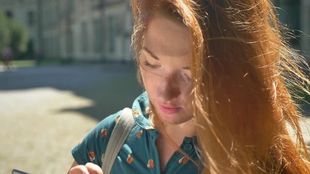 Happy beautiful ginger woman typing on tablet and smiling at camera, standing on street during sunny day