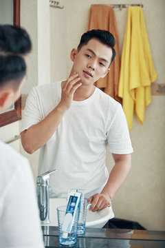 Back View Of Young Asian Man Standing In Front Of Mirror In Bathroom And Exploring Face Skin In Morning