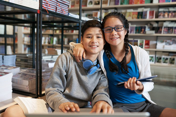 Content girl and boy embracing and holding gadgets while looking at camera in library of school 
