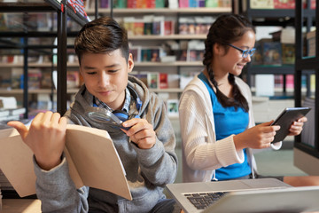 Curious boy exploring book with magnifier and girl using tablet while sitting in school library