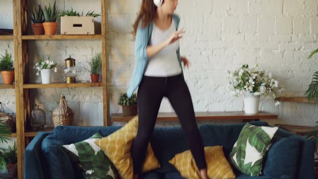 Attractive brunette is having fun at home listening to music through headphones, singing and dancing jumping on sofa. Modern technology and young people concept.