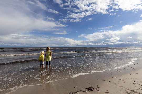 Siblings stand on the shore of a windy sea.
