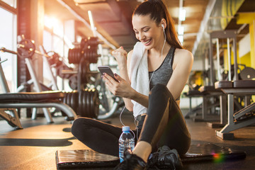 Euphoric young fit woman watching her smart phone while sitting on exercise mat in gym