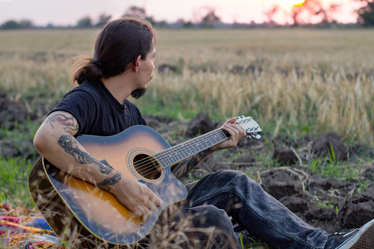 Young tattooed male play on acoustic guitar in summer fields during beautiful sunset	
