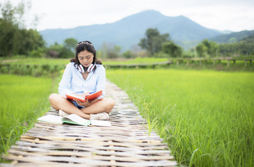 Asian young woman enjoy readbooks on wooden bridge in nature rice farm; her is feeling relax and easy listening music online from mobile device in the meanwhile; Relax and happy concept.