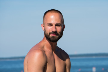 Young handsome athletic adult man with short hair and beard looking into camera while sitting on beautiful tropical beach during hot summer time day. Ocean background scenic view.