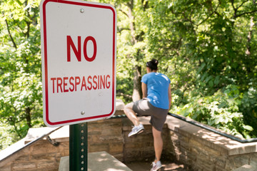 Young woman attempt to jump over railing into restricted area labelled by no trespassing sign. Private property deep woods and forest area illegal to walk past signage - obrazy, fototapety, plakaty