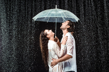 side view of happy couple in white shirts standing under umbrella under raindrops isolated on black