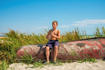 A boy is playing on a black clarinet sitting on an old wooden boat on the beach.